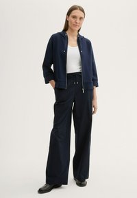 Veste de sport bleu marine avec capuche, fermeture éclair sur le devant et cordon de serrage ; assortie à un pantalon large bleu marine et un débardeur blanc ; texture lisse.