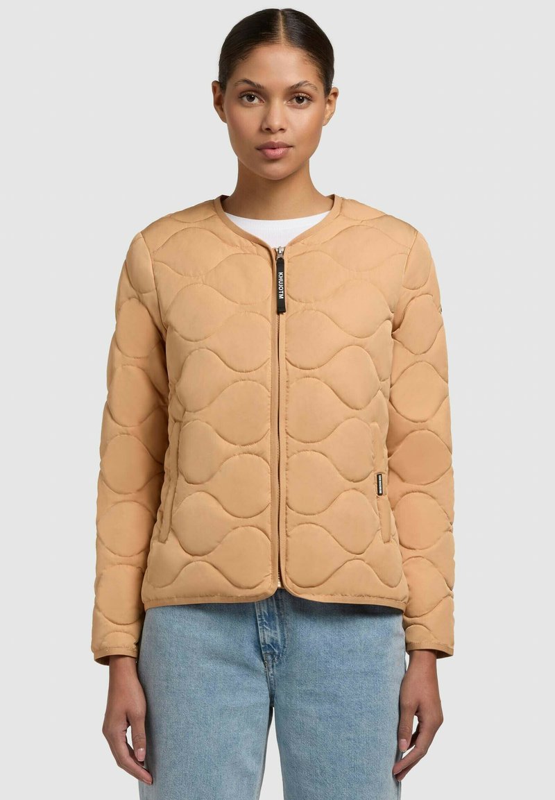 Quiltet beige jakke med rundt halsudskæring, frontlynlås og bølgeformet detaljer. Har lange ærmer og en tætsiddende silhuet.