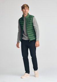 Grüne Steppweste über einem grauen Pullover kombiniert mit dunklen Hosen und beigen Sneakers. Glatter Stoff mit gepolsterter Textur, minimalistisches Design.
