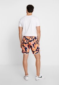 Costumi da bagno da uomo con una base blu navy e vivaci motivi floreali arancioni, vita elasticizzata e bordi bianchi, abbinati a una maglietta bianca.