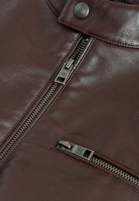 Lähikuva tummanruskean nahkatakin osista, joissa on kaksi metallista vetoketjua ja painonappi.