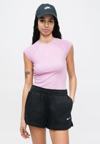 Młoda kobieta w czapce Nike w kolorze czarnym, liliowym bezrękawnym topie i czarnych szortach Nike, z rękami w kieszeniach, stojąca na białym tle.