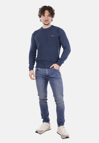 Maglione a coste navy con maniche lunghe, scollatura rotonda e piccolo logo sul petto, abbinato a jeans azzurri chiari e sneaker bianche.