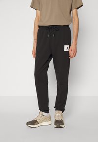 Svarta joggingbyxor med elastisk midja, snörningsdetalj och en vit logotypflik. Kombinerat med bruna träningsskor och en beige t-shirt.