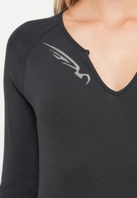 Maglia nera a coste con maniche lunghe e scollatura a V, con un design astratto argentato a forma di ali sul lato sinistro del petto, indossata da una persona con pelle chiara.