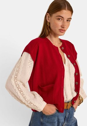 Femme aux longs cheveux bruns portant des boucles d'oreilles créoles dorées, un gilet rouge boutonné sur un chemisier blanc en dentelle, un jean bleu et une ceinture marron.