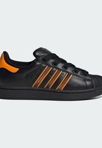 Basket low-top noire avec un talon orange et trois rayures orange et marron sur le côté, dotée de lacets noirs et d'un embout en caoutchouc texturé.