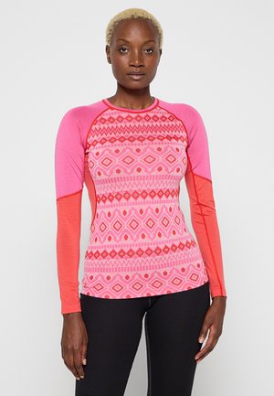 Frau trägt einen taillierten langärmeligen Oberteil mit rosa-rotem Muster und schwarze Hose, steht vor einem schlichten hellen Hintergrund.