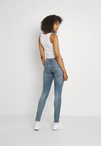 Jeans de denim azul ajustados con una cintura de media altura, bolsillos traseros y un lavado claro, combinados con una blusa sin mangas blanca y zapatillas blancas.