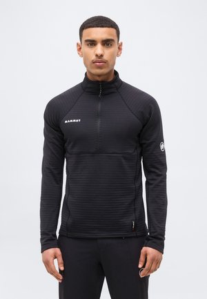 Young man wearing a black Mammut zip-up pullover with long sleeves, standing against a plain white background.