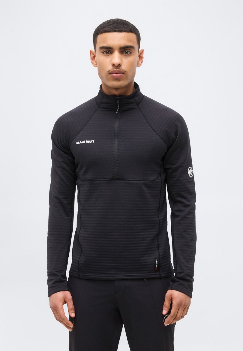 Jeune homme portant un pull à fermeture éclair noir Mammut à manches longues, debout devant un fond blanc uni.