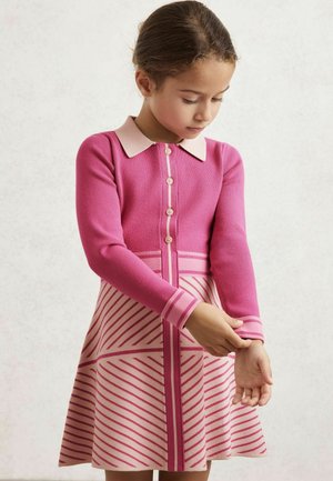 Tütarlaps roosas nööbitud kleitides, millel on triibuline seelik, reguleerib varrukat, vaadates alla lihtsa heleda tausta vastu.