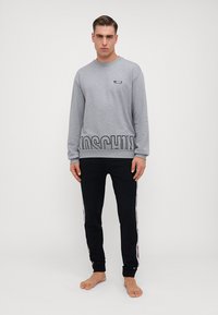 Sivý sweatshirt s dlhým rukávom a čiernym potlačou "MOSCHINO" na leme, kombinovaný s čiernymi teplákmi so bočnými pruhmi a elastickými manžetami na členkoch.