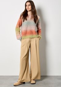 Mehrfarbiger, flauschiger Pullover mit einem Verlauf aus Orange-, Grün- und Blautönen, kombiniert mit beigen, weiten Hosen und hellbeigen Slippern.