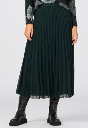 Mujer con falda midi plisada de color verde oscuro, botas negras de cuero hasta la rodilla y un suéter negro con grandes patrones de flores en gris claro.
