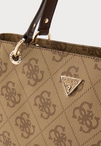 Geantă de mână din țesătură maro cu textură și model în formă de diamant, având un logo triunghiular metalic și accente aurii, precum și o curea maro pentru umăr.