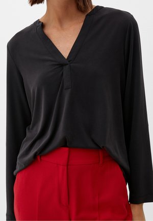 Femme portant un chemisier noir à manches longues col en V, rentré dans un pantalon rouge taille haute, vue des épaules aux hanches.