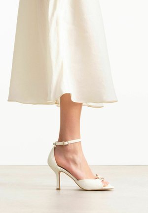 Jambe dans une sandale blanche à talon haut avec bride à la cheville et un petit nœud, partiellement couverte par une jupe fluide couleur crème.