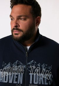 Navy zip-up sweatshirt featuring mountain graphics and white printed text, with a textured fabric and high collar design.