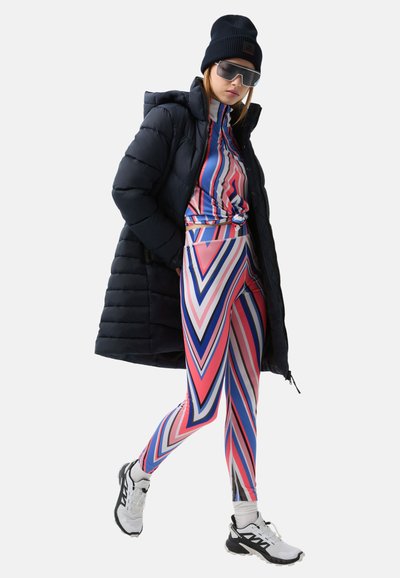 Gepolsterter schwarzer Mantel, bunte legging mit Zickzackmuster und Oberteil in Pink, Blau und Weiß. Accessoires sind eine schwarze Mütze und Sonnenbrillen.