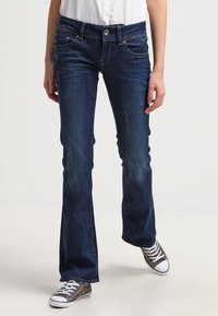 Dark blue flared jeans with a classic five-pocket design, light fading, and a button closure. Worn with gray low-top sneakers.
