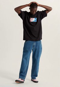 Zwart katoenen T-shirt met een kleurrijk Vans-logo, gecombineerd met blauwe spijkerbroek. De outfit wordt compleet gemaakt met roze en zwarte instappers.