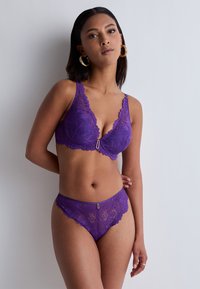 Soutien-gorge en dentelle violette et string assorti avec des bords festonnés, présentant un fermoir décoratif à l'avant. Motif floral complexe sur l'ensemble.