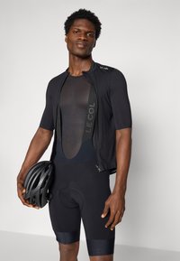 Schwarzes Fahrradtrikot mit kurzen Ärmeln, Netzdetails und einem Frontreißverschluss, kombiniert mit schwarzen Shorts, während er einen schwarzen Helm hält.