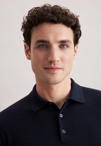 Granatowa koszulka polo z kołnierzykiem, trzema guzikami, miękką teksturą i dopasowanym krojem, noszona przez mężczyznę z kręconymi włosami.