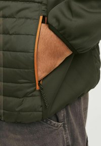 Veste matelassée verte avec un détail de poche zippée orange. Présente une finition en tissu lisse et une section matelassée texturée.