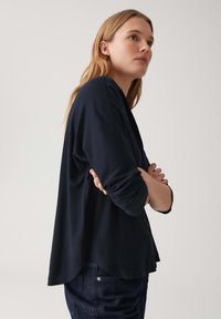 Maglietta a maniche lunghe blu navy con vestibilità rilassata, tessuto morbido, orlo arrotondato e leggero drappeggio. Indossata con jeans scuri, presenta un design semplice senza decorazioni.
