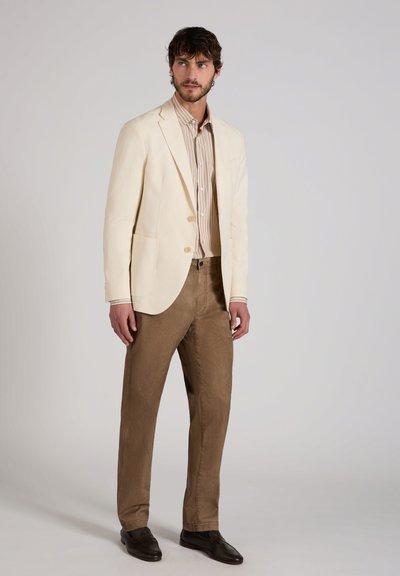 Man draagt een crèmekleurig colbert, beige gestreept overhemd, bruine broek en donkerbruine schoenen, staand tegen een effen grijze achtergrond.