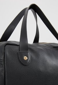 Zwarte leren tas met een gestructureerd oppervlak en gladde zwarte handvatten, bevestigd met gouden klinknagels, tegen een effen lichte achtergrond.