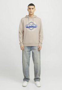 Beige hættebluse med blåt og hvidt grafisk logo, snoretræk i hætten, ribbede ærmer, parret med lyseblå baggy jeans og hvide sneakers.