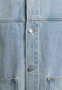 Tissu en denim bleu clair avec deux boutons en métal et deux poches avant, présentant des détails de couture verticale et horizontale.