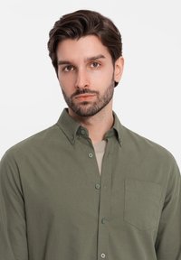 Olijfgroene button-up shirt van zacht materiaal met een puntige kraag, voorzien van een borstzak en bijpassende knopen, gedragen over een licht T-shirt.
