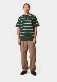 Camiseta de manga corta a rayas verdes con rayas blancas y un pequeño logo. Combinada con pantalones beige y zapatos negros, de pie sobre un fondo sencillo.