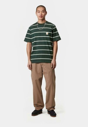 Camiseta de manga corta a rayas verdes con rayas blancas y un pequeño logo. Combinada con pantalones beige y zapatos negros, de pie sobre un fondo sencillo.