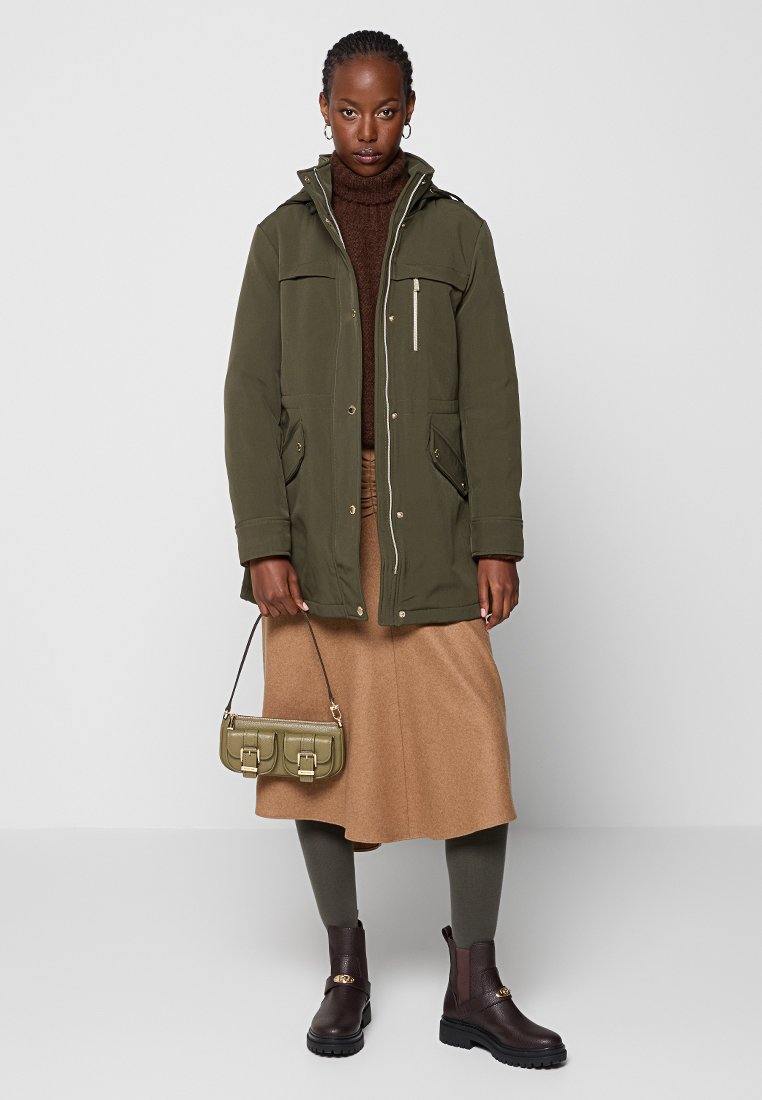 Parka verde oliva con tasche frontali e dettagli con zip, dolcevita marrone, gonna beige, borsa verde e stivaletti marroni. Texture liscia.