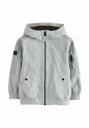 Veste à capuche gris clair avec une finition texturée, fermeture éclair devant, deux poches latérales et des accents en tissu contrastant sur les manches. Patch logo noir visible.