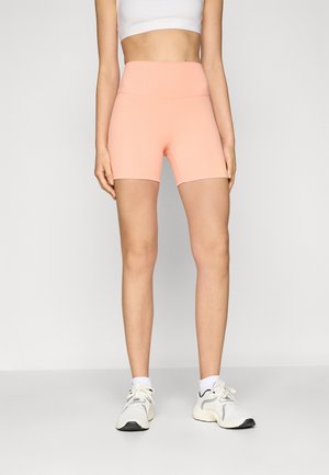 Vrouw draagt hooggetailleerde perzikkleurige fietsshorts, wit cropped topje, witte sokken en witte sneakers, staand tegen een effen lichte achtergrond.