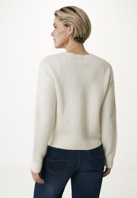 Krämfärgad tröja med texturerad stickning, rundad halsringning och ribbade manschetter, kombinerad med mörkblå jeans. Söm och bakdesign är synlig.