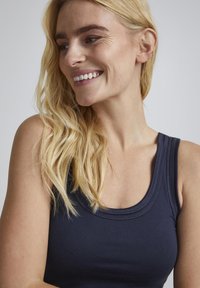 Tumed sinine tank top, millel on ümar kaelus, pehme tekstuur ja kihilised õmbluse detailid. Mudelil on laineline blond juukseviis ja naeratav ilme.