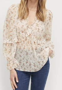Blusa floreale in tessuto leggero e trasparente con bordi arricciati, scollatura a V e maniche lunghe a sbuffo; presenta piccoli motivi floreali su fondo crema.