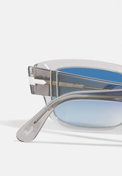 Óculos de sol com armação cinza transparente e lentes espelhadas azuis. Apresenta detalhes em prata e o logotipo "Persol" em relevo na haste.
