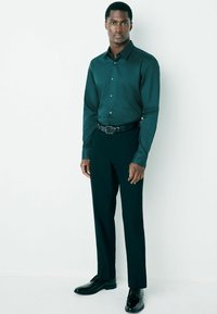Dunkelgrünes Hemd mit Knopfleiste und glatter Textur, kombiniert mit schwarzen Hosen und glänzenden schwarzen Businessschuhen. Klassischer Kragen und taillierte Passform.