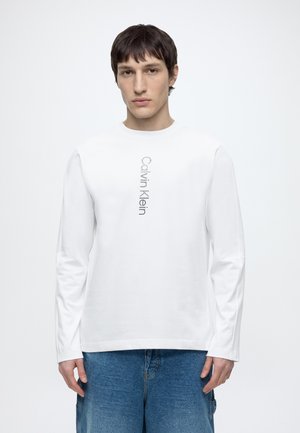 Mladý muž s krátkými tmavými vlasy, oblečený v bílém tričku Calvin Klein s dlouhým rukávem a modrými džínami, stojící před jednobarevným pozadím.