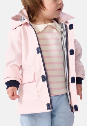 Roze waterdichte jas met capuchon, marineblauwe accenten en drukknoopsluiting. Voorzien van een voorzak en gestreepte voering. Gecombineerd met een gestreept shirt.