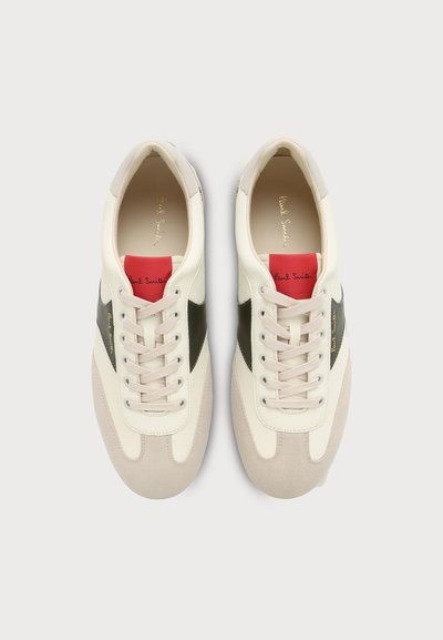 Paire de baskets blanches et beiges avec des panneaux latéraux noirs et des étiquettes rouges sur la languette, vues de dessus sur un fond clair.