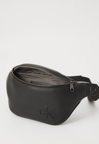 Borsa in pelle nera a vita con zip aperta, foderata internamente in grigio, logo "cK" in rilievo e tracolla regolabile.
